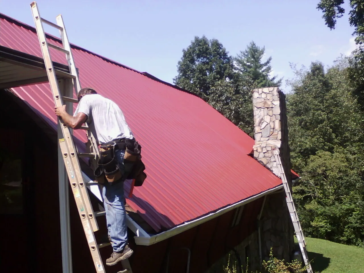 Roofer on a ladder beside a red metal roof and stone chimney — Soffit & Fascia Repair by Roofing Brothers in Hay Springs